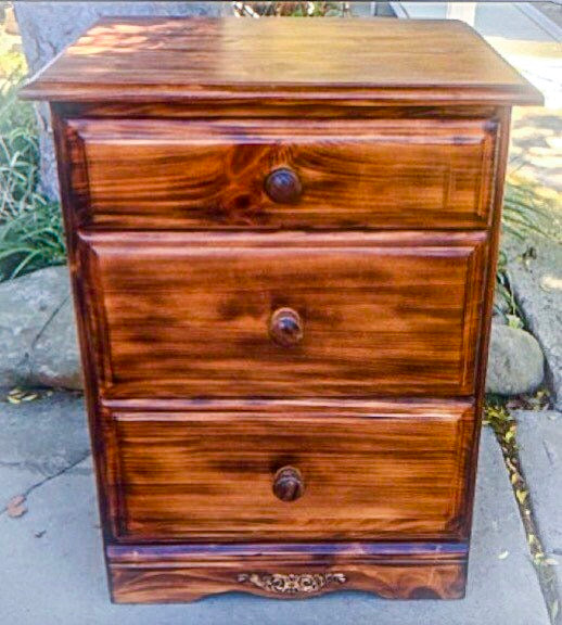 A solid wood, vintage nightstand with three drawers. The upcycled nightstand has been done in a burnt wood finish.