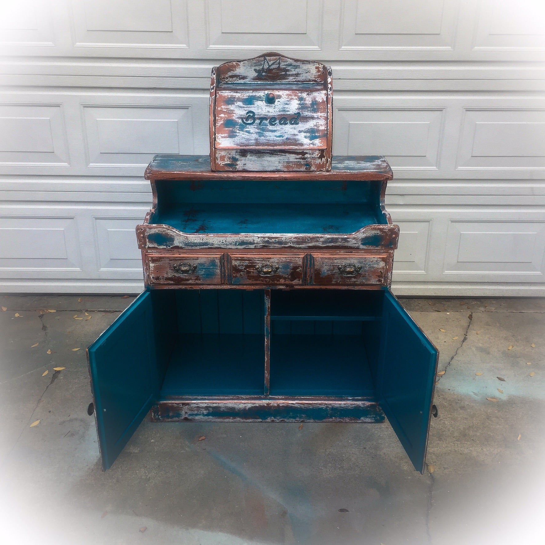 A piece of vintage furniture, repurposed into a coffee bar, painted in distressed blue, shown with it’s cabinet area open.
