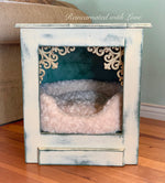 A distressed white, end table, with wrought iron accents & a pet bed in the large cubby underneath.