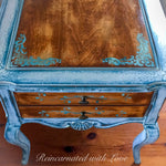 A close up of the front portion on an end table, in stained wood with white & blue, damask accents & patina, drawer pulls.