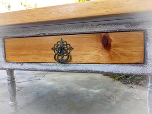 Close up of a knotted, pine drawer, with an antique drawer pull, on a distressed white table.