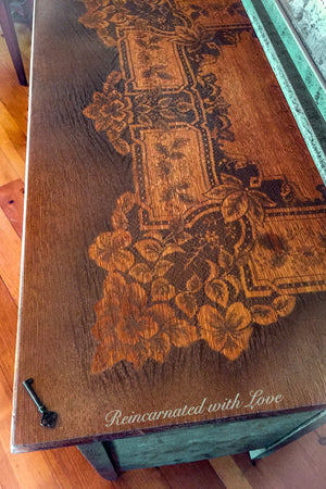 Close up of the tabletop on a farmhouse style, dressing table, with green, patina trim, stained wood & lace accents.