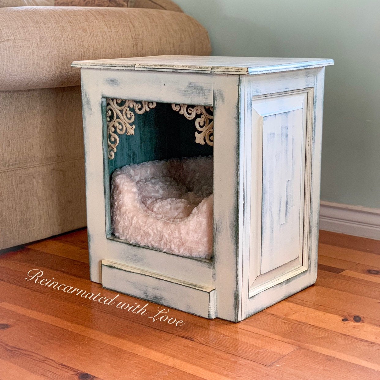 An upcycled, end table with the front, cabinet door removed & a pet bed added, painted in an off white with green undertones.