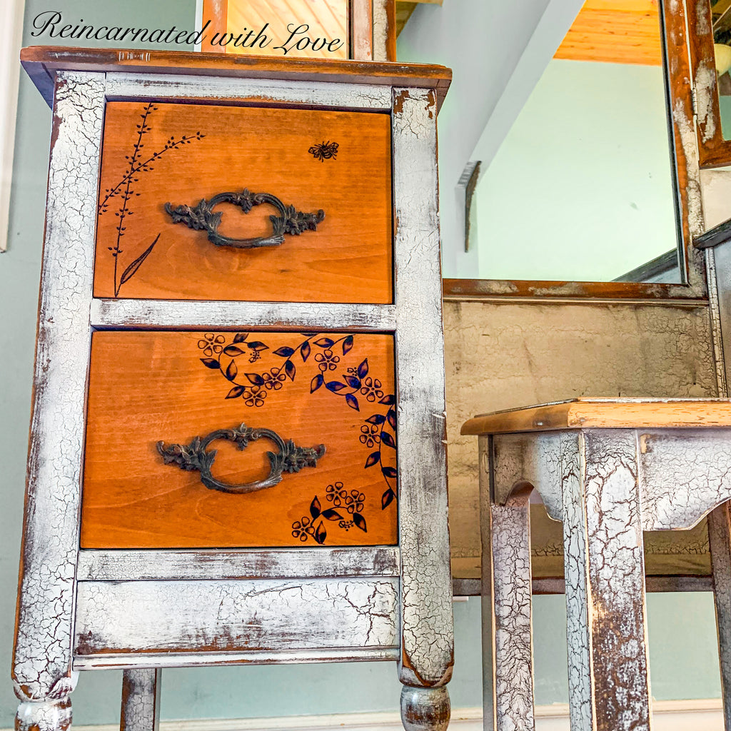 An antique vanity table in distressed white with stained wood tabletop & drawer faces. The vanity has burnt wood accents.