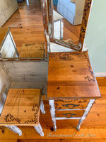 A makeup table that has burnt wood floral accents. Painted in distressed white with stained wood tabletop & drawer faces.